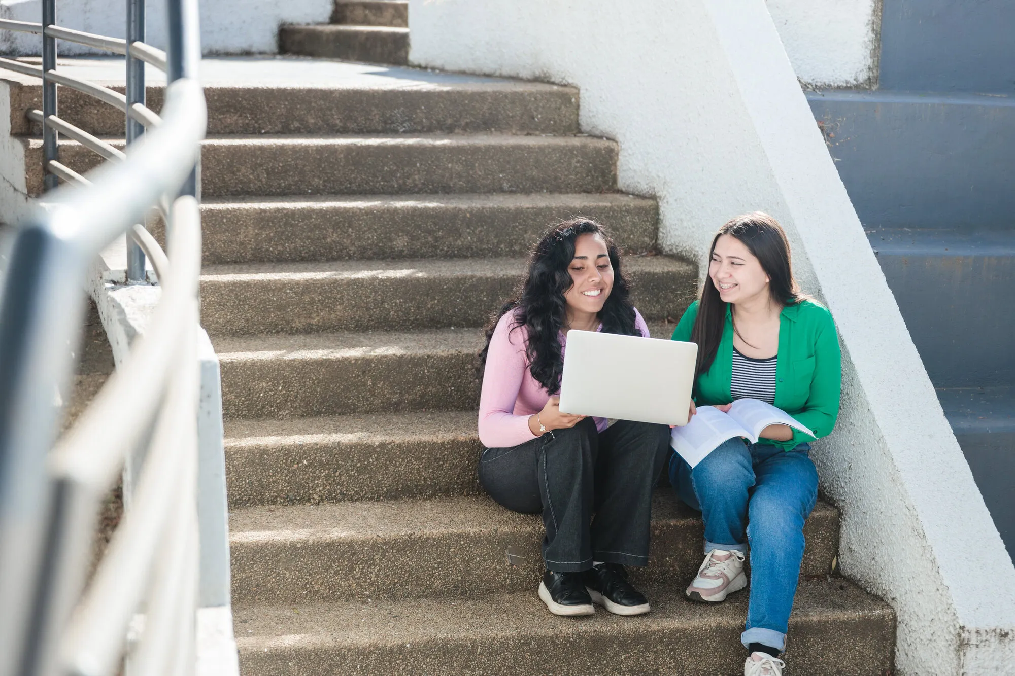 Front view of two latin student women studying with a laptop and book, sitting in staircase campus schoolyard